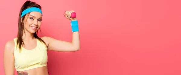 Attractive sportswoman training with dumbbell while smiling at camera on pink background — Stock Photo