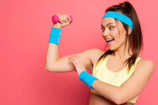 Aufgeregte Sportlerin berührt Bizeps beim Training mit Hantel auf rosa Hintergrund — Stockfoto