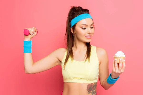 Young cheerful sportswoman holding dumbbell and looking at delicious cupcake on pink background — Stock Photo