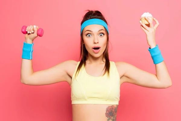 Schockierte junge Sportlerin mit Hantel und leckerem Cupcake vor der Kamera auf rosa Hintergrund — Stockfoto