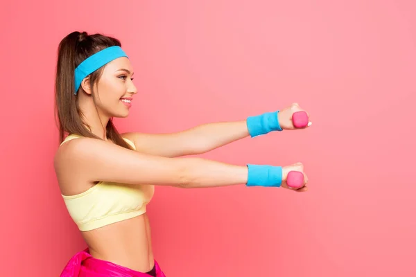 Side view of smiling young sportswoman training with dumbbells on pink background — Stock Photo