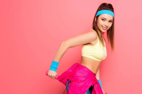 Hermosa joven deportista de entrenamiento con mancuerna mientras sonríe a la cámara sobre fondo rosa - foto de stock