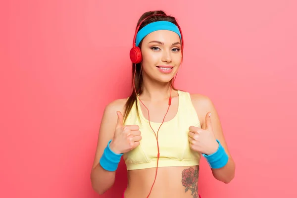 Happy young sportswoman in headphones showing thumbs up on pink background — Stock Photo