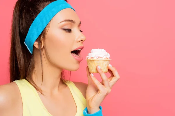 Young sportswoman going to eat delicious cupcake with whipped cream isolated on pink — Stock Photo