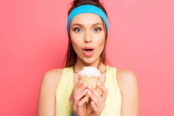 Возбужденная спортсменка держит вкусный кекс, глядя на камеру на розовом фоне — стоковое фото