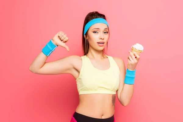 Young sportswoman showing disapproval gesture while holding cupcake on pink background — Stock Photo