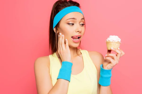 Verwirrte Sportlerin hält leckeren Cupcake in der Hand und leckt sich die Lippen isoliert auf rosa — Stockfoto