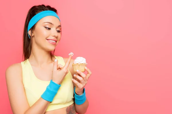 Allegra sportiva con panna montata sul dito che tiene delizioso cupcake isolato sul rosa — Foto stock