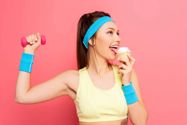Felice sportiva che tiene i manubri e mangia deliziosi cupcake su sfondo rosa — Foto stock