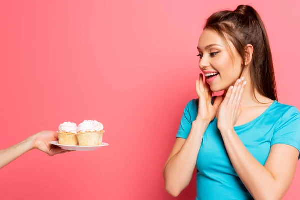 Schockierte Sportlerin hält Hand nahe Gesicht nahe weiblicher Hand mit Teller mit leckeren Cupcakes auf rosa Hintergrund — Stockfoto