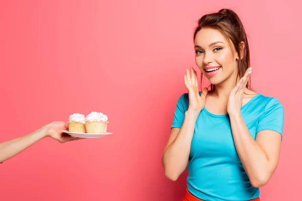 Aufgeregte Sportlerin hält Hand nahe Gesicht nahe weiblicher Hand mit Teller mit leckeren Cupcakes auf rosa Hintergrund — Stockfoto