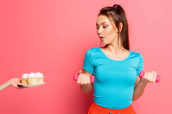 Überraschte Sportlerin beim Training mit Hanteln in der Nähe weiblicher Hand mit einem Teller leckerer Cupcakes auf rosa Hintergrund — Stockfoto