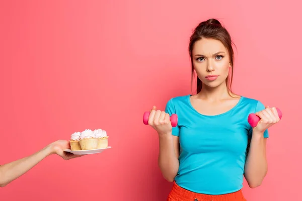 Deportista confiado haciendo ejercicio con pesas cerca de la mano femenina con plato de deliciosos cupcakes sobre fondo rosa - foto de stock