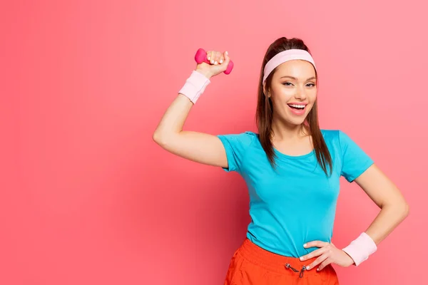 Happy sportswoman exercising with dumbbell while holding hand on hip on pink background — Stock Photo