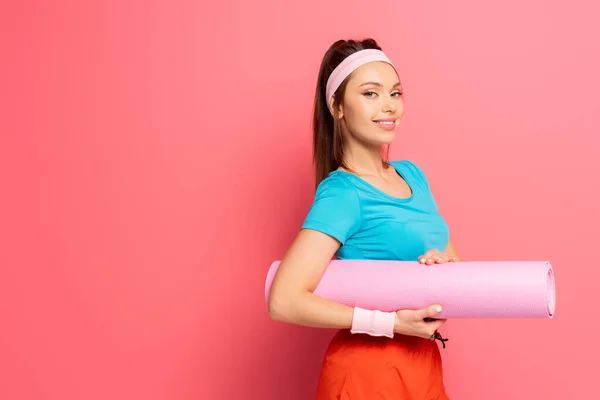 Attraktive, glückliche Sportlerin mit Fitnessmatte, während sie auf rosa Hintergrund in die Kamera lächelt — Stockfoto