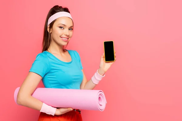 Deportista sonriente sosteniendo la alfombra de fitness y mostrando el teléfono inteligente con pantalla en blanco aislado en rosa - foto de stock