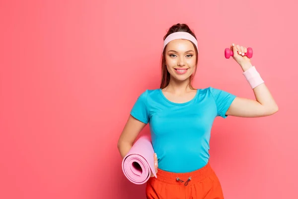 Deportista feliz sosteniendo mancuerna y alfombra de fitness mientras sonríe a la cámara sobre fondo rosa - foto de stock