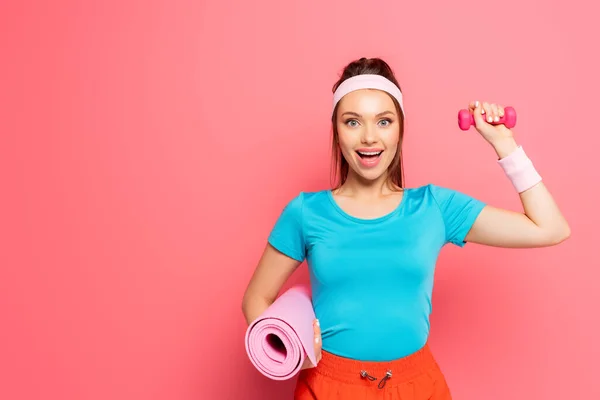 Sportiva eccitata che tiene i manubri e il tappetino fitness mentre guarda la fotocamera su sfondo rosa — Foto stock