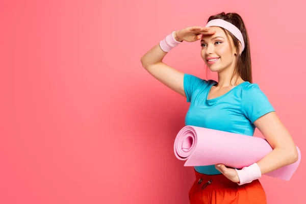 Lächelnde Sportlerin, die wegschaut, während sie eine Fitnessmatte auf rosa Hintergrund hält — Stockfoto