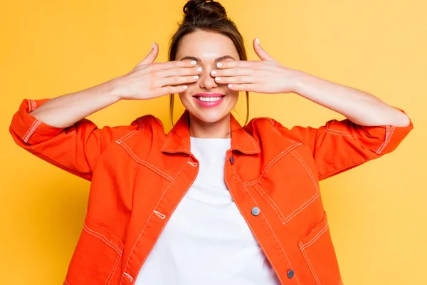 Chica sonriente cubriendo los ojos con las manos sobre fondo amarillo - foto de stock