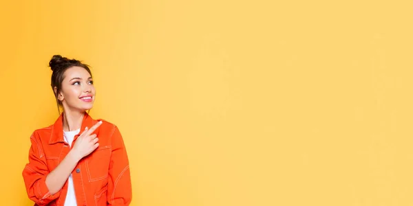 Panoramic shot of attractive smiling woman looking away and pointing with finger isolated on yellow — Stock Photo