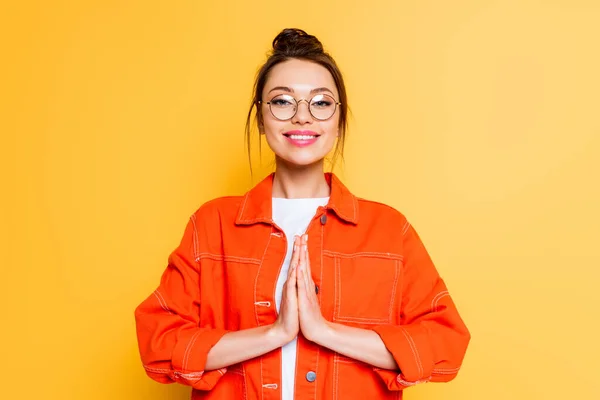 Smiling student in eyeglasses standing with praying hands isolated on yellow — Stock Photo
