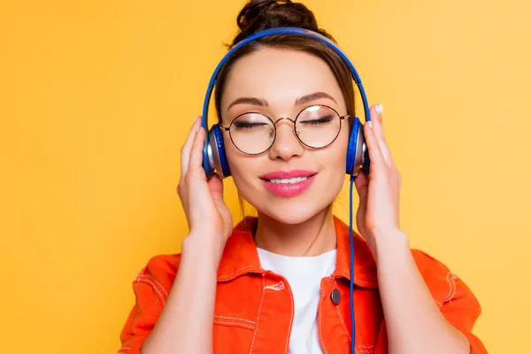 Smiling student in eyeglasses touching headphones while standing with closed eyes isolated on yellow — Stock Photo