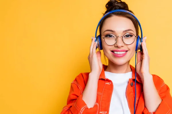 Happy student in eyeglasses touching headphones while smiling a camera isolated on yellow — Stock Photo