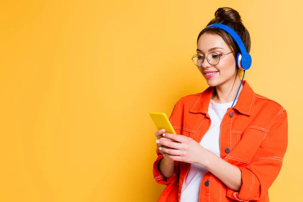 Estudiante sonriente en auriculares charlando en el teléfono inteligente sobre fondo amarillo - foto de stock