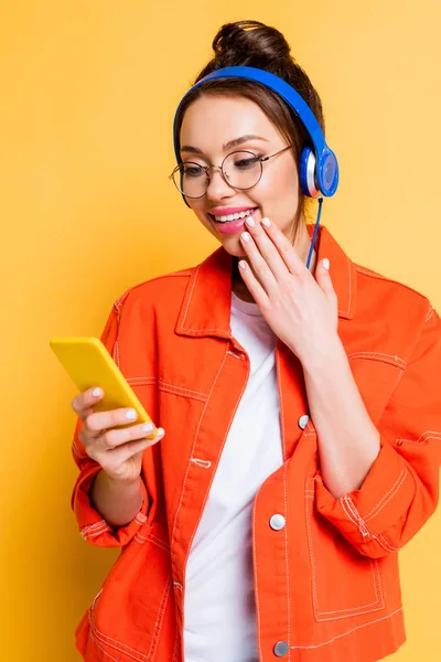 Estudiante sorprendido en auriculares charlando en el teléfono inteligente y tocando la cara en el fondo amarillo - foto de stock