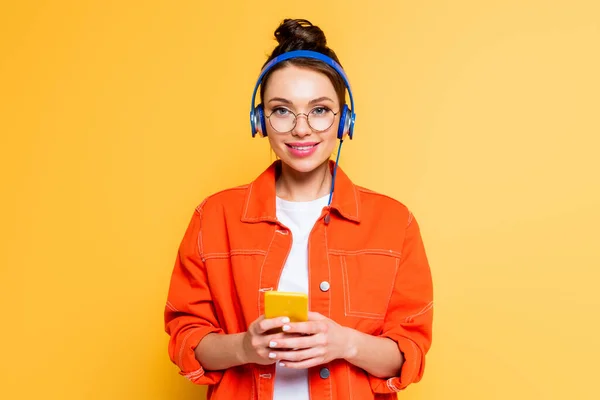 Estudiante sonriente en auriculares sonriendo a la cámara mientras sostiene el teléfono inteligente aislado en amarillo — Stock Photo
