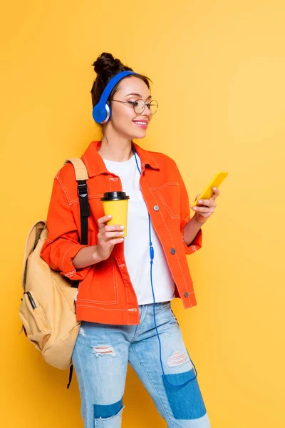 Estudiante alegre en auriculares usando teléfono inteligente mientras sostiene el café para ir sobre fondo amarillo - foto de stock