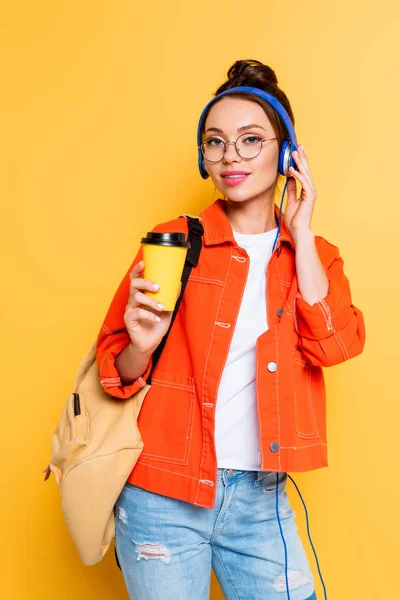 Atractivo estudiante tocando auriculares mientras sostiene el café para ir sobre fondo amarillo - foto de stock