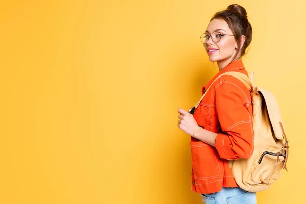 Attraktive, lächelnde Studentin mit Rucksack, die in die Kamera auf gelbem Hintergrund blickt — Stockfoto