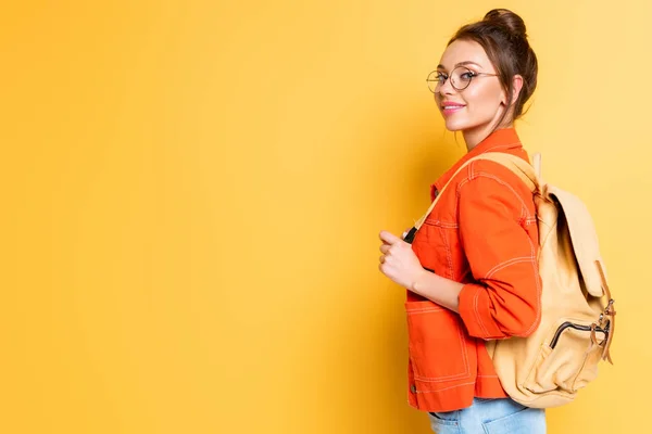 Atractiva estudiante sonriente con mochila mirando a la cámara sobre fondo amarillo - foto de stock