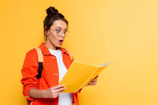 Shocked student in glasses looking at copy book isolated on yellow — Stock Photo