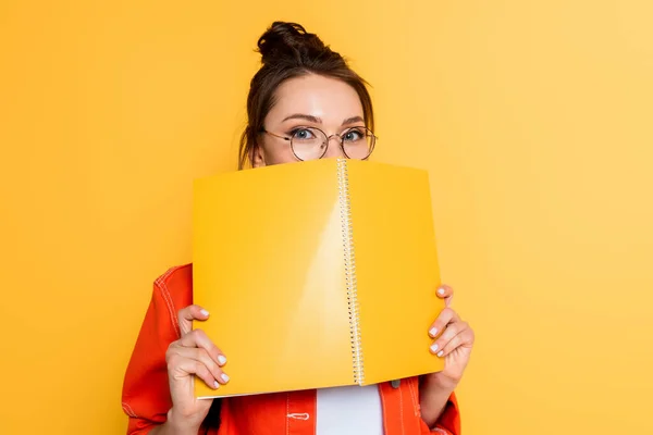 Jovem estudante em óculos obscurecendo rosto com livro de cópia enquanto olha para a câmera isolada no amarelo — Fotografia de Stock