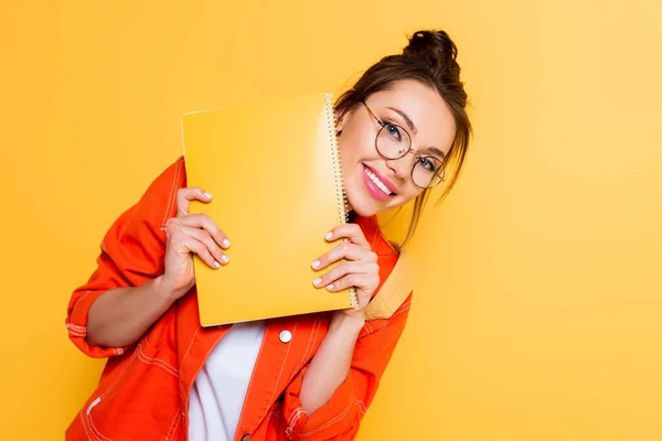 Estudiante alegre en gafas mirando a la cámara mientras sostiene cuaderno amarillo aislado en amarillo - foto de stock