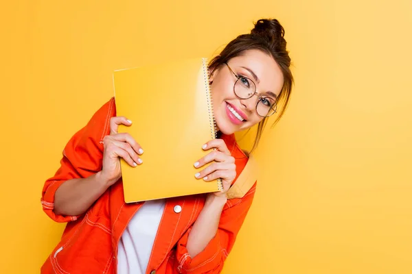 Cheerful student in glasses looking at camera while holding yellow notebook isolated on yellow — Stock Photo