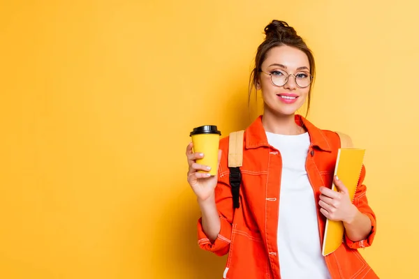 Atractivo, estudiante feliz sosteniendo café para ir y portátil mientras sonríe a la cámara en el fondo amarillo - foto de stock