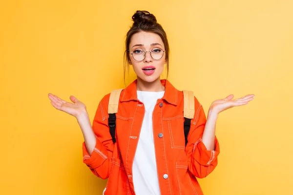 Étudiant confus dans les lunettes montrant geste haussant les épaules tout en regardant la caméra sur fond jaune — Photo de stock