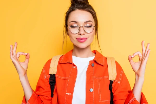 Estudiante sonriente en gafas de pie en pose de meditación con los ojos cerrados aislados en amarillo - foto de stock