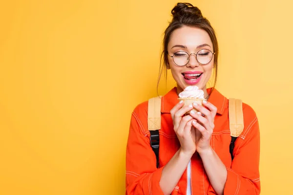 Счастливая студентка с закрытыми глазами, засовывая губы, держа вкусный кекс на желтом фоне — стоковое фото