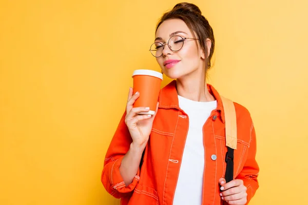 Joyful student in eyeglasses enjoying flavor of coffee with closed eyes isolated on yellow — Stock Photo