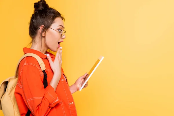 Вид сбоку шокированного студента, держащего руку возле рта, держа цифровую планшет изолированным на желтом — стоковое фото