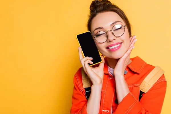 Estudiante feliz celebración de teléfono inteligente con pantalla en blanco con los ojos cerrados sobre fondo amarillo - foto de stock