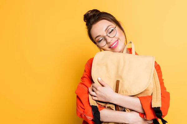 Estudiante feliz en gafas que sostiene la mochila con los ojos cerrados sobre fondo amarillo - foto de stock