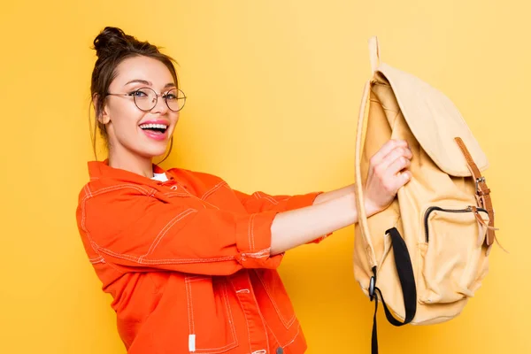 Estudiante feliz sosteniendo la mochila mientras sonríe a la cámara en el fondo amarillo - foto de stock