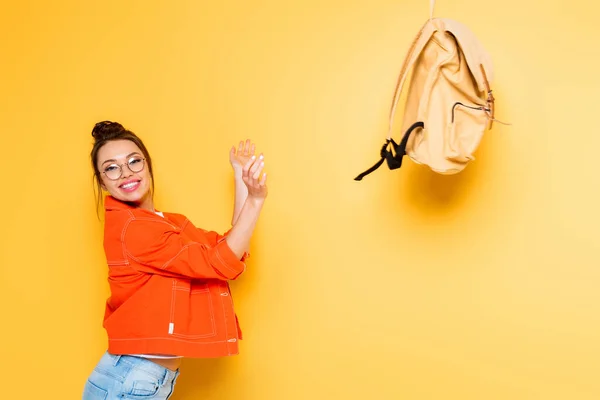 Excited student throwing backpack while smiling at camera on yellow background — Stock Photo