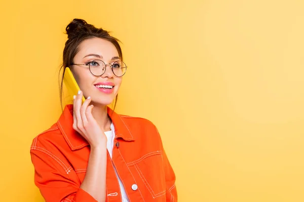 Cheerful student in eyeglasses talking on smartphone while looking away isolated on yellow — Stock Photo